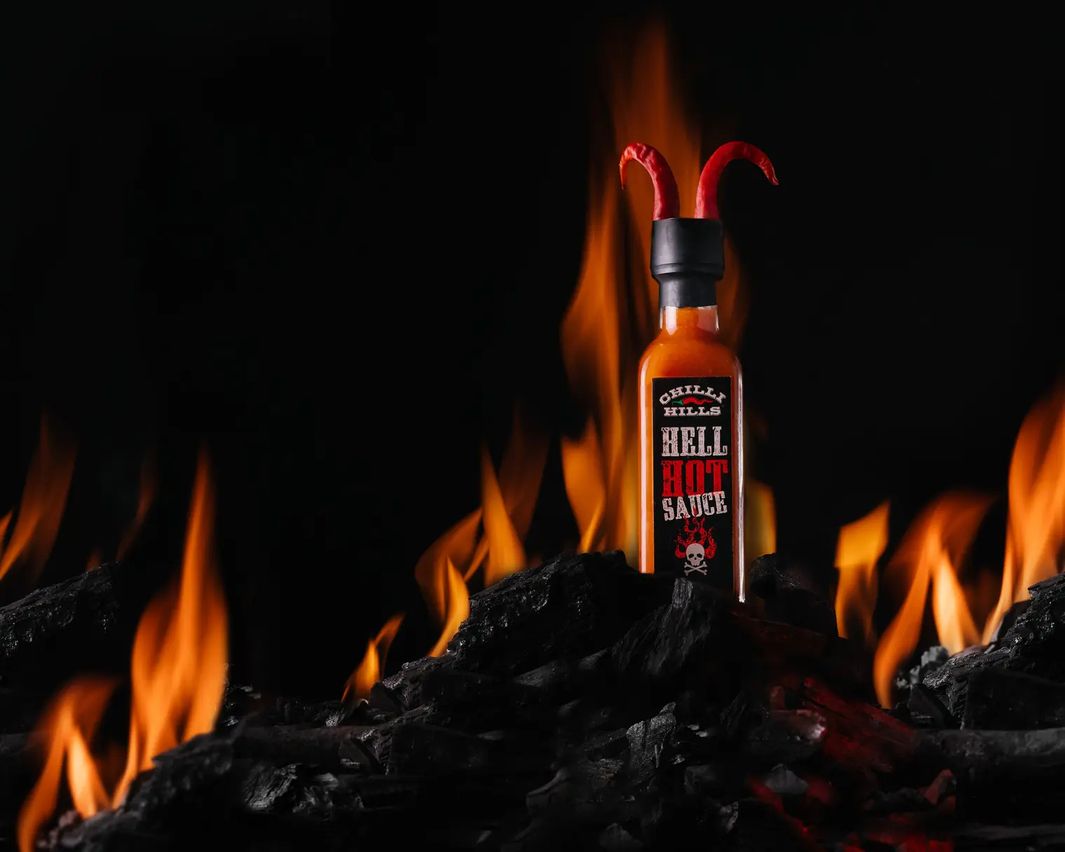Chilli Hills Hell Hot Sauce върху горещи въглени. Буркан с горещ сос Chilli Hills Hell Hot Sauce стои върху горящи въглища. Зад нея гори огън. В буркана има рогца от люти чушки, които трябва да подскажат на зрителя – сосът е дяволски опасен.