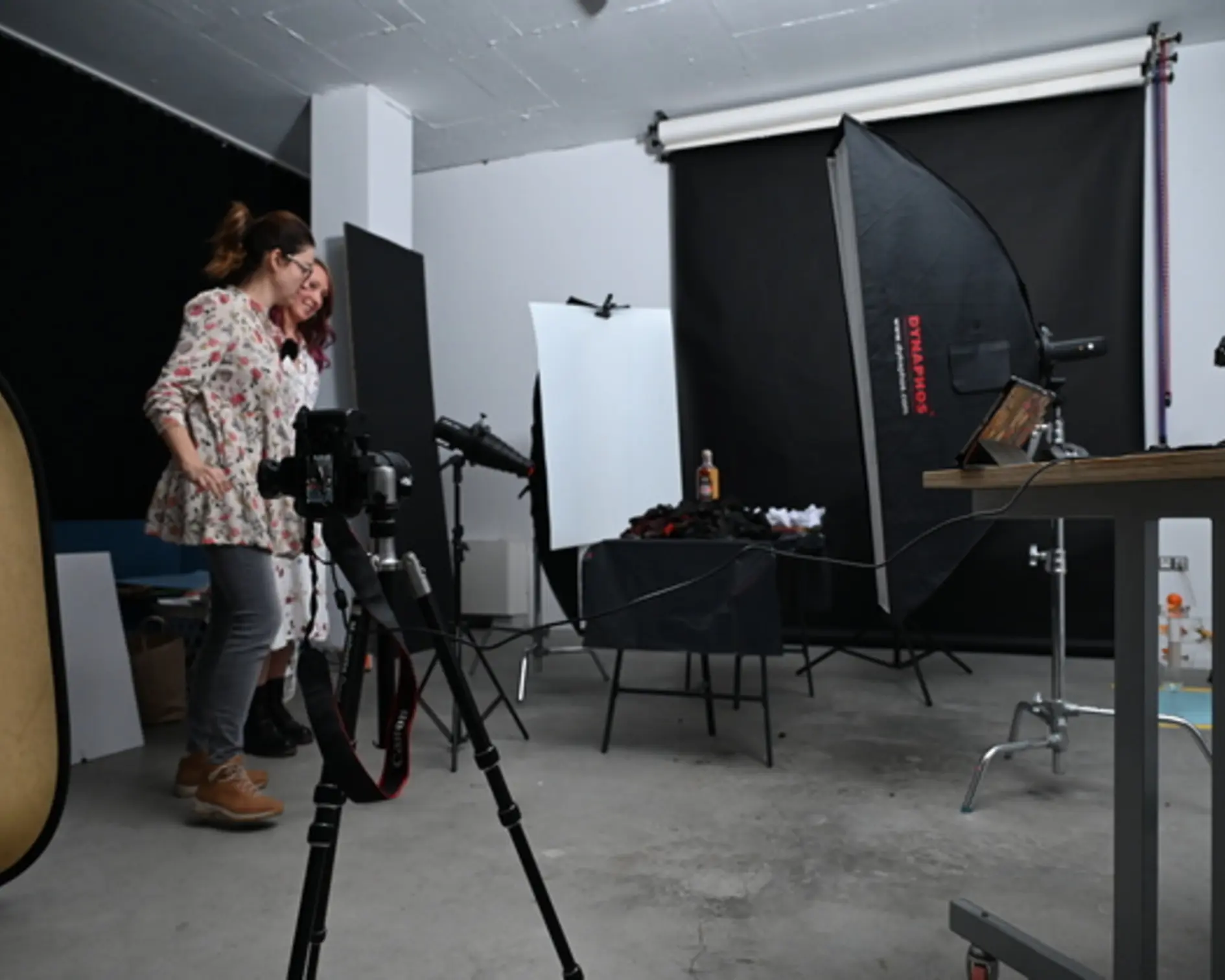 Диляна Гергова и Олга Бабицкая в студио Dynaphos. Две жени стоят в студио Dynaphos и обсъждат как да заснемат следващата композиция. Расставлены стрипбоксы и на масата има обект за снимане.
