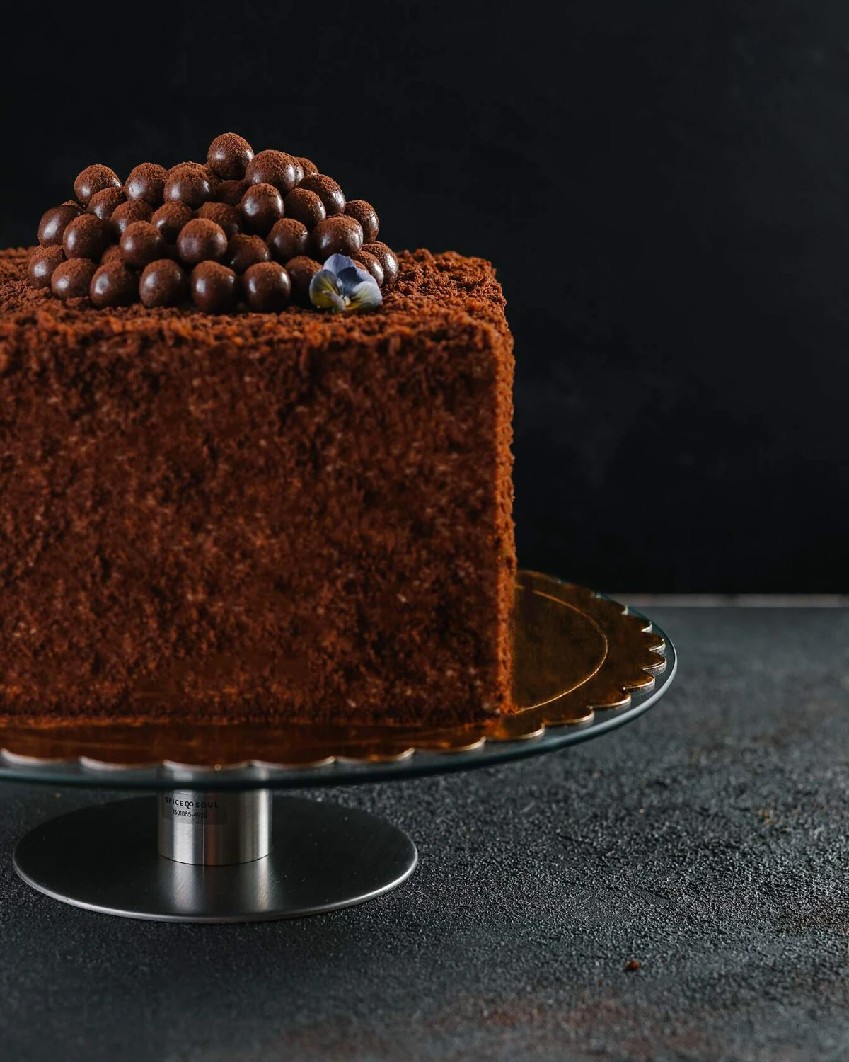 Бисквитена шоколадово медена торта. На тъмен фон има квадратна торта върху поставка за торта. Декорирана е с шоколадови топчета и виолетови цветя.