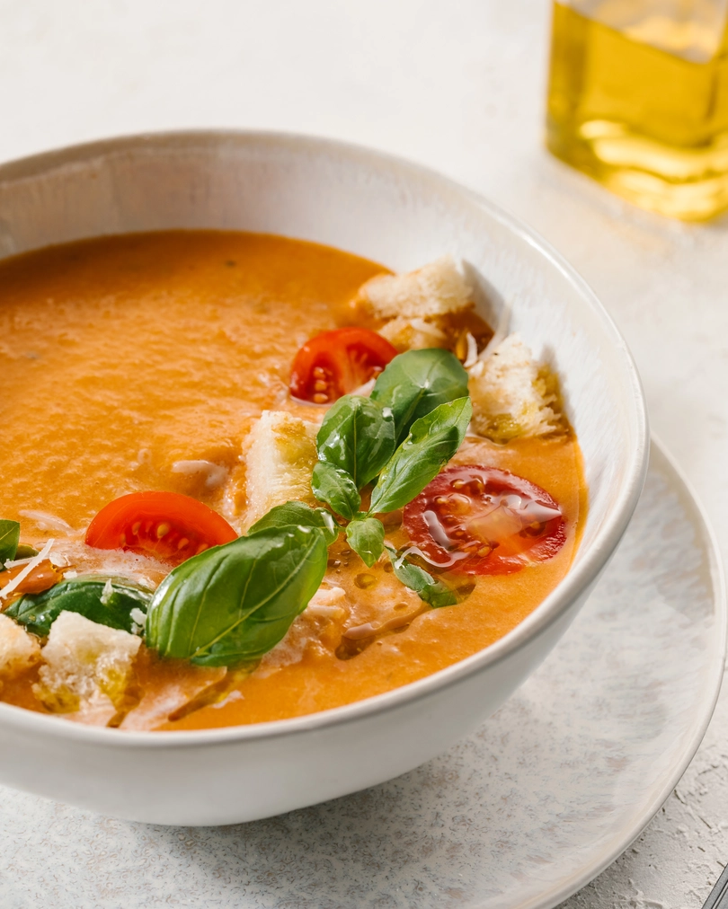 Супа от печени домати. В чинията е показана доматена супа отблизо. Гарнира се с крутони от бял хляб, зехтин и чери домати. На заден план на снимката има съд за зехтин.
