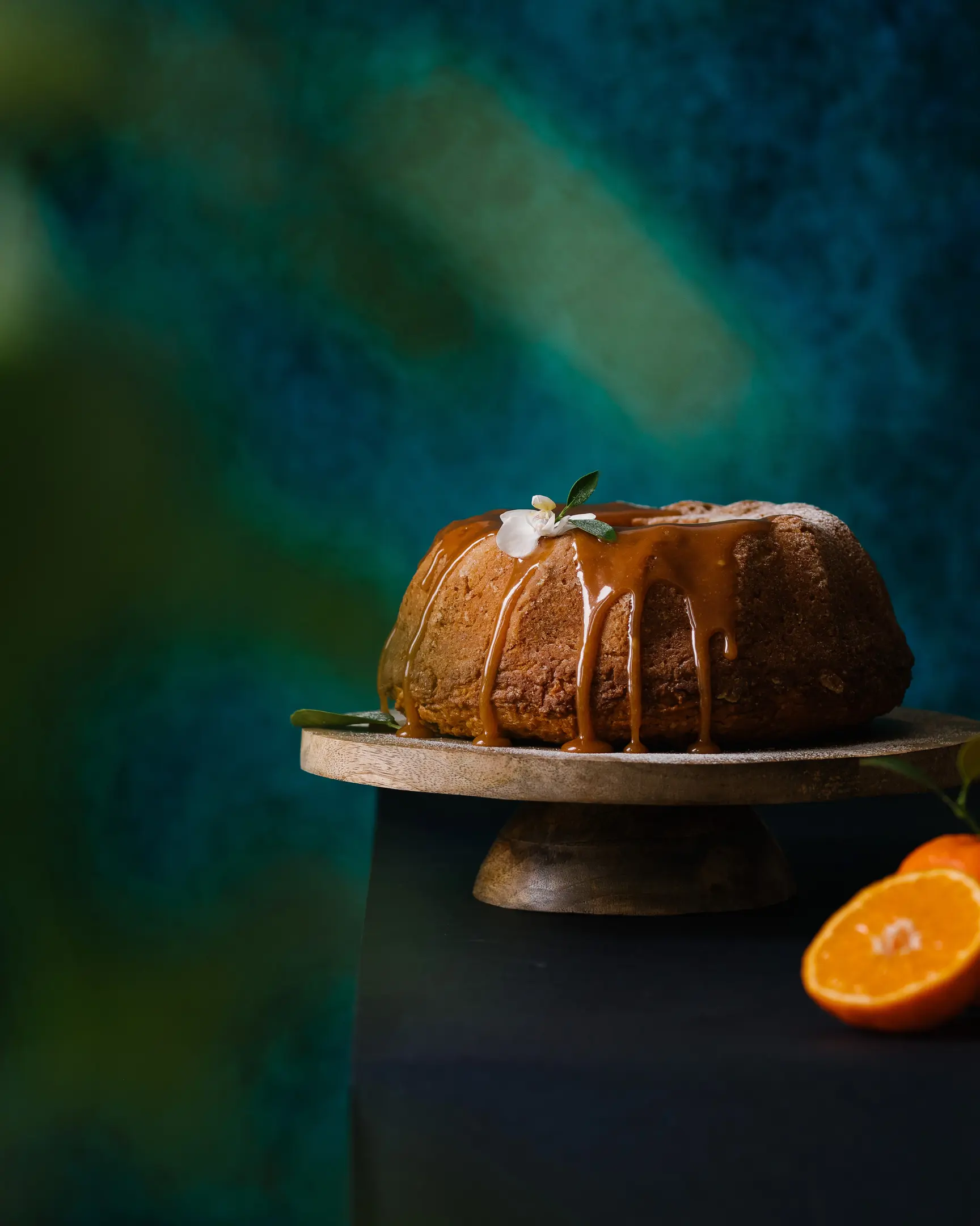На тюркоазен фон има оранжев кекс. На тюркоазен фон има оранжев кекс върху дървена поставка за торта. Покрито е с карамел. Зелените листа са на преден план на рамката, което придава дълбочина на рамката.