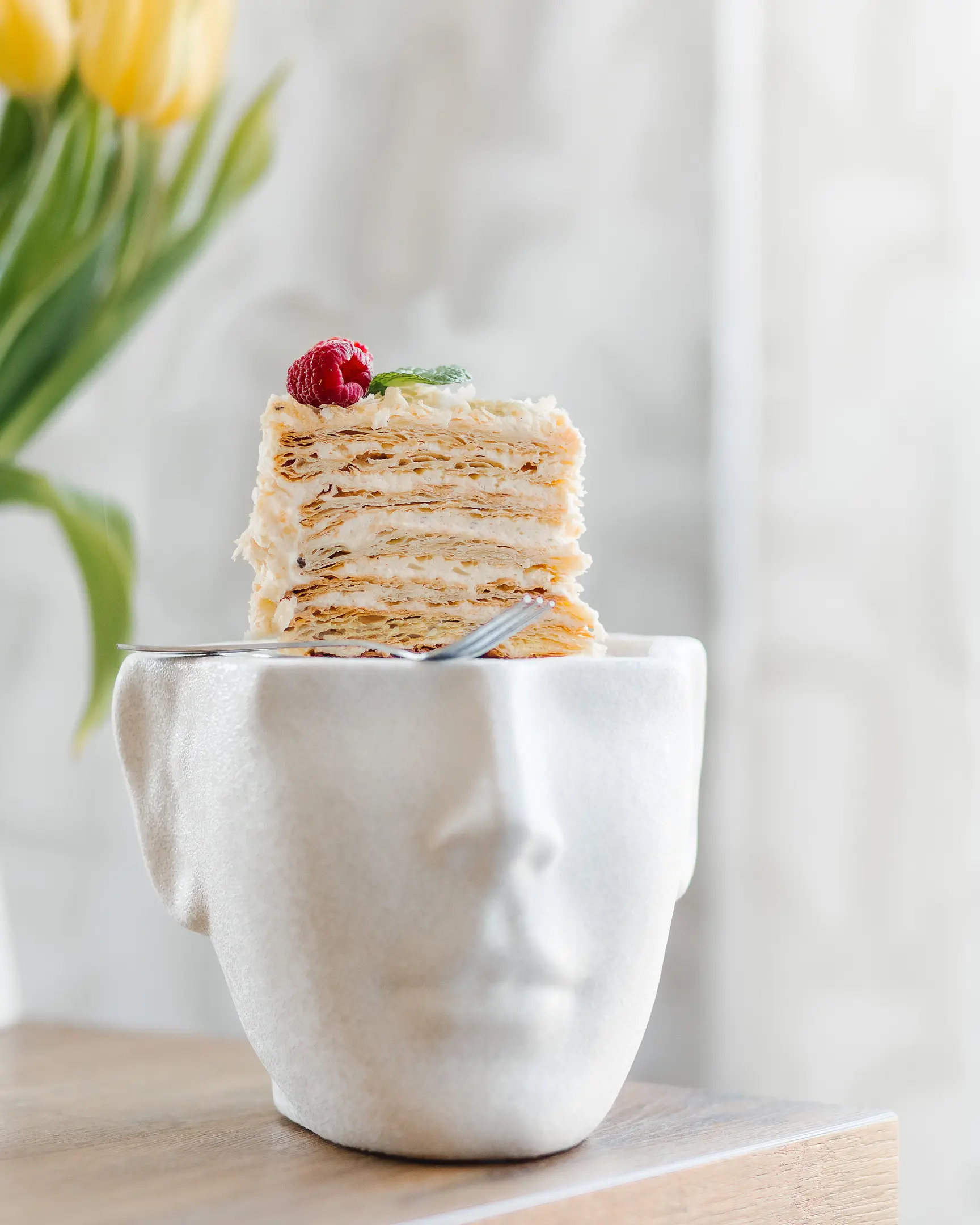 Парче торта Наполеон. Парче бутер торта Наполеон стои върху чиния под формата на човешка глава. Тортата е на масата. На заден план има цветя в буркан