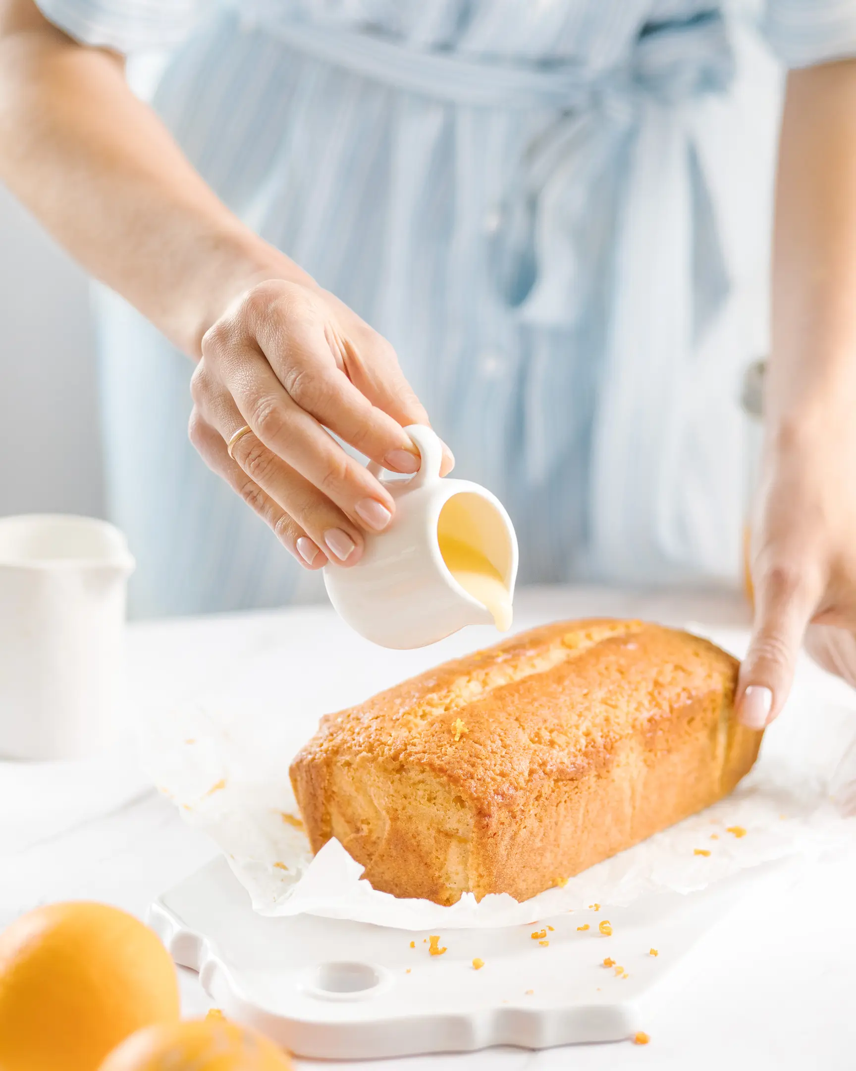Жена излива портокалова глазура върху кекс. Жена в синя рокля излива портокалова глазура върху кекс. Кексът стои върху керамична поставка. На преден план се виждат портокали, което подчертава, че кексът е направен на базата на портокали.