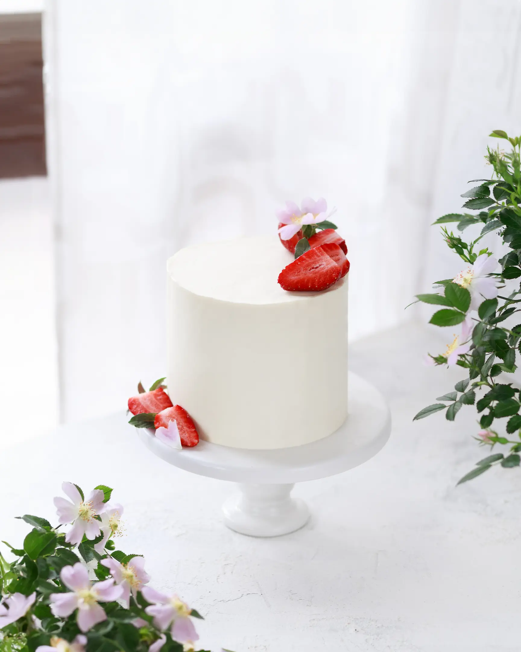 На светъл фон медена торта покрита с бял крем. На светъл фон срещу прозореца стои торта, украсена с горски плодове. Отстрани на рамката се виждат цветове от шипка.